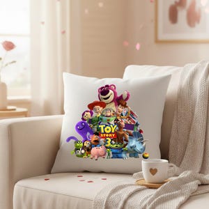 May include: White square pillow featuring a colorful graphic of characters from the animated film "Toy Story 3." The design includes Woody, Buzz Lightyear, and other beloved characters. The pillow is placed on a beige sofa.