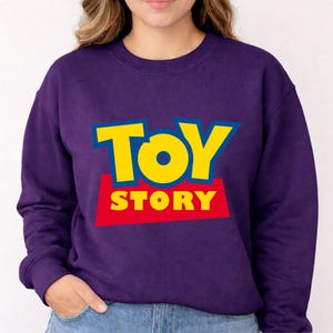 May include: A purple sweatshirt featuring the words "TOY STORY" in yellow and red block letters. The sweatshirt is a crew neck style and appears to be made of a soft material. The design is a reference to the animated movie.