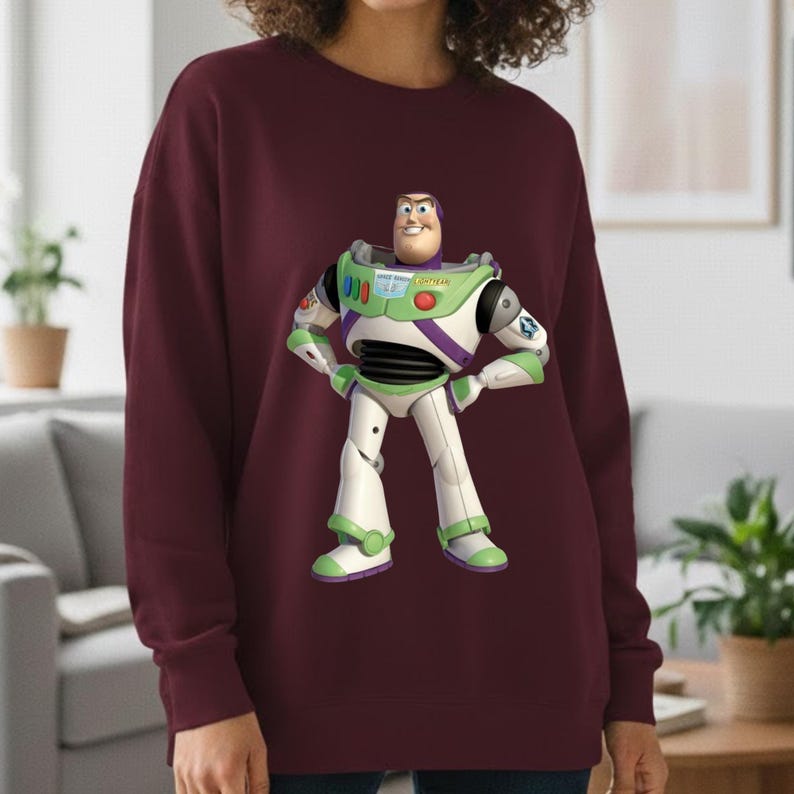May include: A maroon sweatshirt featuring a colorful graphic of Buzz Lightyear from Toy Story. The character is depicted in his signature white, green, and purple spacesuit, with his arms on his hips. The sweatshirt is a solid color.