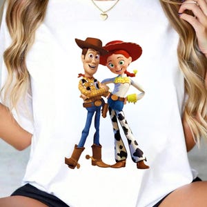 May include: White t-shirt featuring a colorful graphic of Woody and Jessie from Toy Story. Woody is wearing a brown cowboy hat, a yellow plaid shirt, blue jeans, and brown boots. Jessie is wearing a red hat, a white shirt, blue jeans, and cow-print pants.