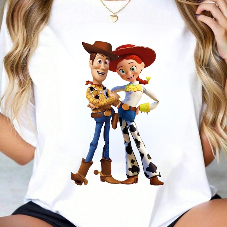 May include: White t-shirt featuring a colorful graphic of Woody and Jessie from Toy Story. Woody is wearing a brown cowboy hat, a yellow plaid shirt, blue jeans, and brown boots. Jessie is wearing a red hat, a white shirt, blue jeans, and cow-print pants.