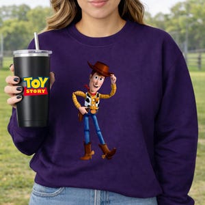 May include: A purple sweatshirt featuring a graphic of Woody from Toy Story. A black tumbler with the Toy Story logo is held in the hand. The tumbler has a silver straw and lid.