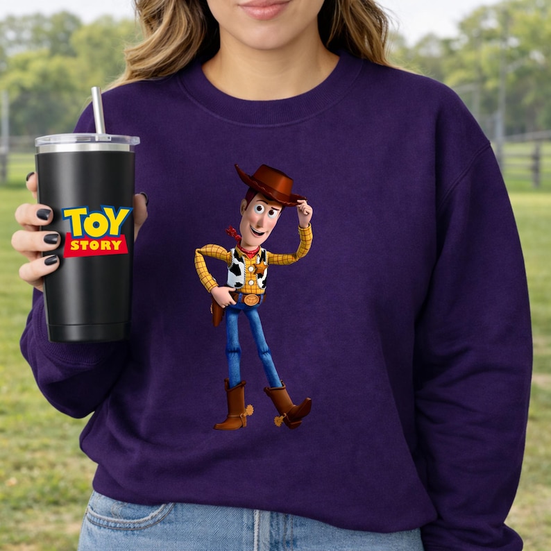 May include: A purple sweatshirt featuring a graphic of Woody from Toy Story. A black tumbler with the Toy Story logo is held in the hand. The tumbler has a silver straw and lid.