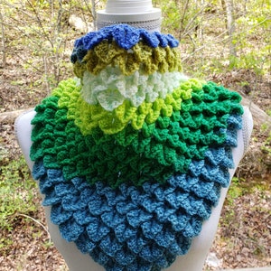 May include: A crocheted cowl with a dragon scale pattern in shades of blue, green, and yellow. The cowl is displayed on a white mannequin.