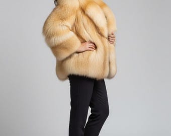 May include: A luxurious, golden-toned fur coat with a voluminous silhouette. The coat features a plush texture and is paired with black trousers and ankle boots. The model is smiling, posing in front of a neutral background.