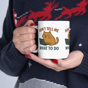 Puede incluir: Taza de cerámica blanca con un gato de dibujos animados con expresión enfadada trabajando en un portátil. La taza tiene el texto "DON'T TELL ME WHAT TO DO" impreso. La taza es sostenida por una persona que lleva un jersey azul oscuro.