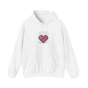 Puede incluir: Sudadera con capucha blanca con bolsillo delantero y cordón. La parte delantera presenta un diseño de corazón pixelado con un mando de juego y el texto "LEVEL UP YOUR GIFTS!" en verde y rosa.