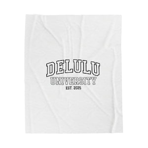 Puede incluir: Manta blanca con el texto "DELULU UNIVERSITY EST. 2025" en negro, con letras arqueadas. La manta tiene un patrón sutil y texturizado en su superficie.