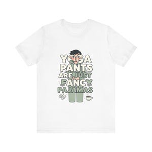 Könnte beinhalten: Weißes T-Shirt mit dem Text "YOGA PANTS ARE JUST FANCY PAJAMAS" in einer verspielten Schriftart. Das Design zeigt eine Cartoonfigur in grüner Hose, Hausschuhen und einer Kaffeetasse, was eine humorvolle und lässige Stimmung erzeugt.