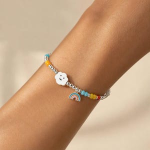 May include: A beaded bracelet featuring small metallic silver spheres, a white cloud charm with a simple black smiley face, and a small colorful rainbow pendant. The design includes interspersed blue, yellow, and red beads along the silver chain.