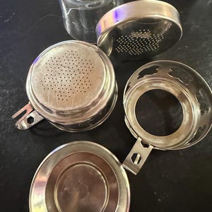 Puede incluir: Un juego de infusor de t&eacute; desmontado con una taza de vidrio transparente y varios componentes met&aacute;licos plateados. El juego incluye una cesta de infusi&oacute;n perforada, una tapa y un soporte, todo dise&ntilde;ado para t&eacute; de hojas sueltas.