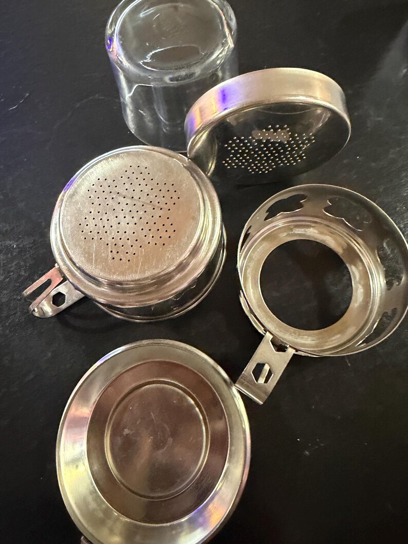 Puede incluir: Un juego de infusor de t&eacute; desmontado con una taza de vidrio transparente y varios componentes met&aacute;licos plateados. El juego incluye una cesta de infusi&oacute;n perforada, una tapa y un soporte, todo dise&ntilde;ado para t&eacute; de hojas sueltas.