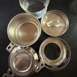 Puede incluir: Un juego de infusor de t&eacute; de plata desmontado con una taza de vidrio transparente. El juego incluye un infusor perforado, una tapa, una base y una bandeja de goteo. Los componentes est&aacute;n dispuestos sobre una superficie oscura.