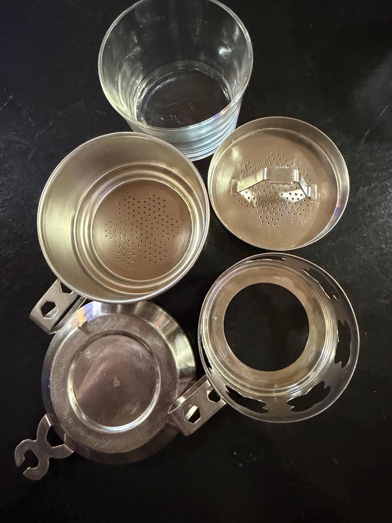 Puede incluir: Un juego de infusor de t&eacute; de plata desmontado con una taza de vidrio transparente. El juego incluye un infusor perforado, una tapa, una base y una bandeja de goteo. Los componentes est&aacute;n dispuestos sobre una superficie oscura.