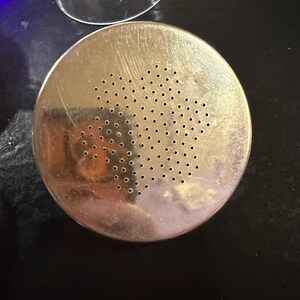 Puede incluir: Un colador met&aacute;lico redondo de color plateado con numerosos agujeros peque&ntilde;os. El colador tiene un aspecto ligeramente desgastado y est&aacute; colocado sobre una superficie oscura. Un objeto de vidrio transparente es parcialmente visible en el fondo.