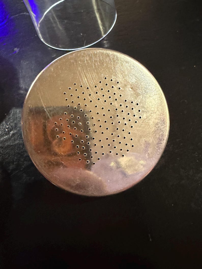 Puede incluir: Un colador met&aacute;lico redondo de color plateado con numerosos agujeros peque&ntilde;os. El colador tiene un aspecto ligeramente desgastado y est&aacute; colocado sobre una superficie oscura. Un objeto de vidrio transparente es parcialmente visible en el fondo.