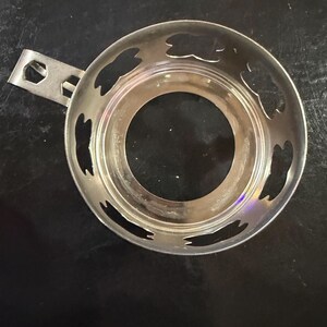 Puede incluir: Un calentador de t&eacute; plateado con asa y recortes decorativos. El calentador circular tiene una abertura central y un asa peque&ntilde;a para facilitar su uso. Los recortes tienen forma de hoja.