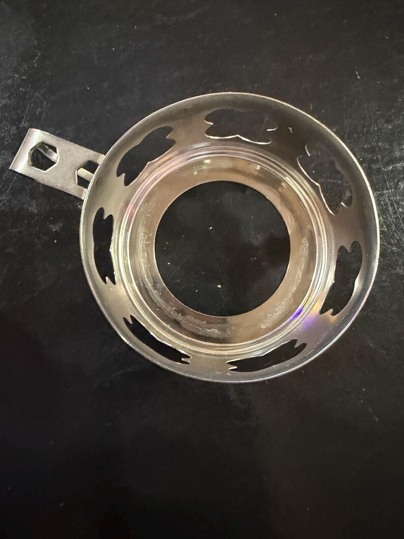 Puede incluir: Un calentador de t&eacute; plateado con asa y recortes decorativos. El calentador circular tiene una abertura central y un asa peque&ntilde;a para facilitar su uso. Los recortes tienen forma de hoja.