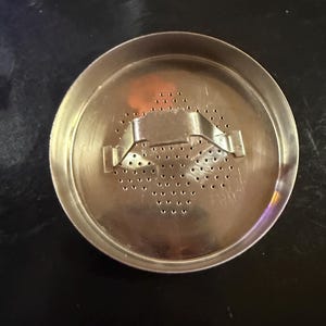 Puede incluir: Un infusor de t&eacute; met&aacute;lico circular con un dise&ntilde;o perforado. El infusor tiene un asa con bisagras y numerosos agujeros peque&ntilde;os para infusionar hojas de t&eacute;. El metal tiene una superficie brillante y reflectante.