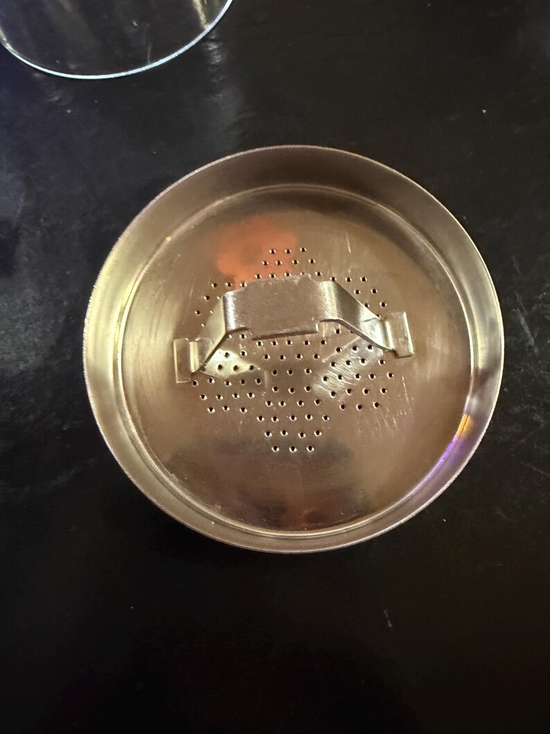 Puede incluir: Un infusor de t&eacute; met&aacute;lico circular con un dise&ntilde;o perforado. El infusor tiene un asa con bisagras y numerosos agujeros peque&ntilde;os para infusionar hojas de t&eacute;. El metal tiene una superficie brillante y reflectante.