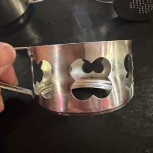 Puede incluir: Un portavasos plateado con asa y formas decorativas de tr&eacute;bol recortadas. El portavasos es de metal y tiene un acabado brillante. Varios otros portavasos son visibles en el fondo.