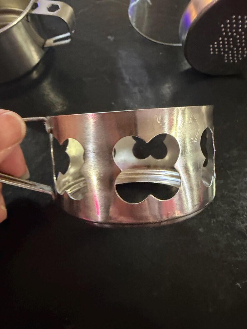 Puede incluir: Un portavasos plateado con asa y formas decorativas de tr&eacute;bol recortadas. El portavasos es de metal y tiene un acabado brillante. Varios otros portavasos son visibles en el fondo.