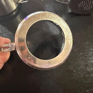 Puede incluir: Primer plano de un anillo de taza de caf&eacute; de acero inoxidable con asa. El anillo es circular con un agujero en el centro. Otras partes de la taza de caf&eacute; de acero inoxidable son visibles en el fondo.