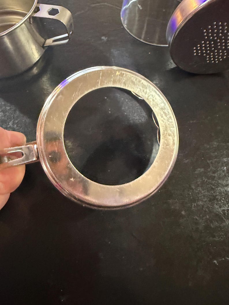 Puede incluir: Primer plano de un anillo de taza de caf&eacute; de acero inoxidable con asa. El anillo es circular con un agujero en el centro. Otras partes de la taza de caf&eacute; de acero inoxidable son visibles en el fondo.