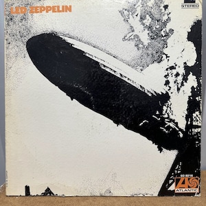 Puede incluir: Una portada de disco de vinilo en blanco y negro con el texto "Led Zeppelin" en letras naranjas. La portada presenta una imagen en blanco y negro de un dirigible volando sobre una ciudad. La discográfica es Atlantic Records y el número de disco es SD 8216.