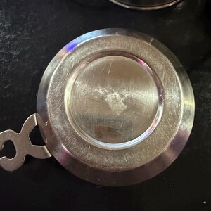 Puede incluir: Una tapa de acero inoxidable con asa y dise&ntilde;o circular. La tapa tiene una superficie brillante y reflectante y probablemente sea para una taza o un recipiente peque&ntilde;o. El asa tiene una forma &uacute;nica.