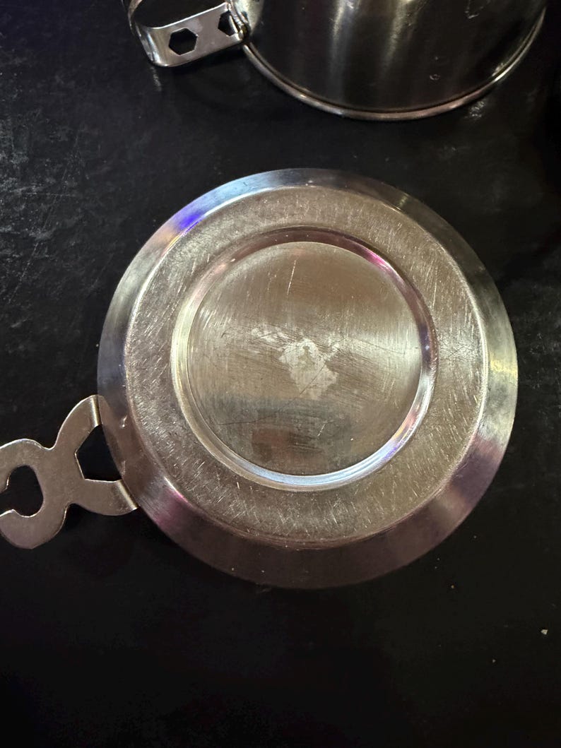 Puede incluir: Una tapa de acero inoxidable con asa y dise&ntilde;o circular. La tapa tiene una superficie brillante y reflectante y probablemente sea para una taza o un recipiente peque&ntilde;o. El asa tiene una forma &uacute;nica.