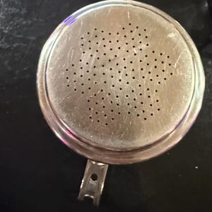 Puede incluir: Un colador redondo de metal plateado con numerosos agujeros peque&ntilde;os. El colador tiene un asa y un borde ligeramente elevado. La superficie muestra signos de desgaste, con ara&ntilde;azos visibles. Dise&ntilde;ado para colar l&iacute;quidos o s&oacute;lidos.