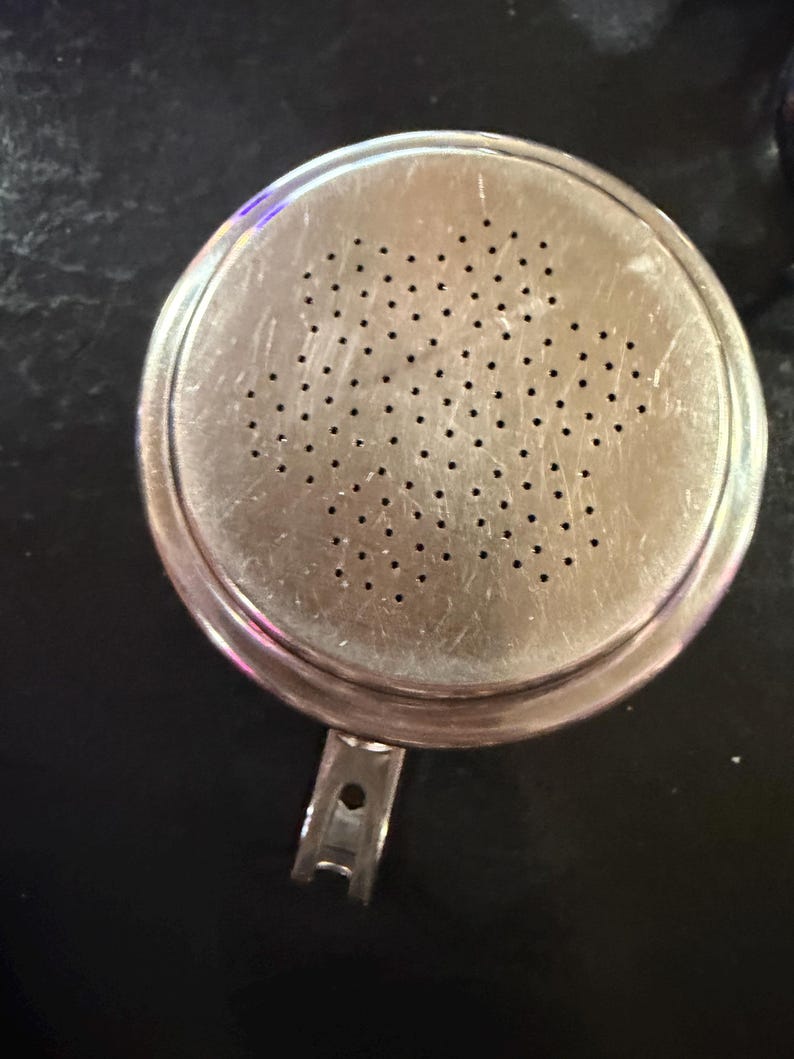 Puede incluir: Un colador redondo de metal plateado con numerosos agujeros peque&ntilde;os. El colador tiene un asa y un borde ligeramente elevado. La superficie muestra signos de desgaste, con ara&ntilde;azos visibles. Dise&ntilde;ado para colar l&iacute;quidos o s&oacute;lidos.