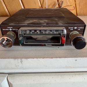 May include: Classic car radio with a dark brown fascia and silver detailing. It has knobs for balance, volume, and tuning, with FM and AM band selection. The radio face shows frequency numbers and the brand name.