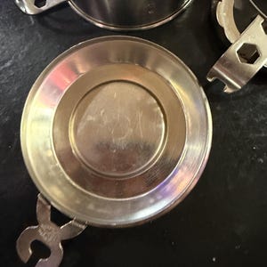 Puede incluir: Primer plano de varios componentes de taza de t&eacute; de metal plateado brillante. Los componentes incluyen una tapa, una taza y un asa. Los componentes est&aacute;n sobre una superficie oscura. El asa tiene un dise&ntilde;o &uacute;nico.