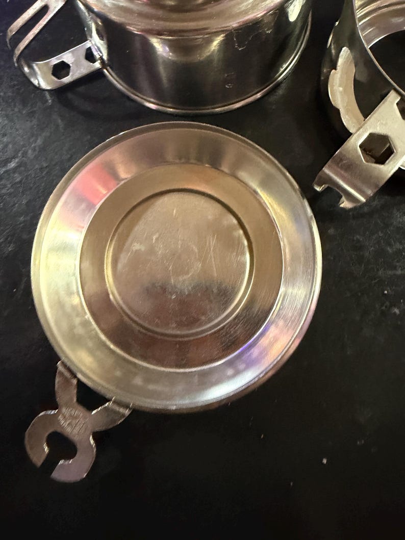 Puede incluir: Primer plano de varios componentes de taza de t&eacute; de metal plateado brillante. Los componentes incluyen una tapa, una taza y un asa. Los componentes est&aacute;n sobre una superficie oscura. El asa tiene un dise&ntilde;o &uacute;nico.