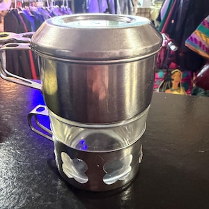 Puede incluir: Una cafetera vintage de acero inoxidable con una jarra de cristal. La cafetera tiene tapa, asa y una base decorativa con dise&ntilde;os recortados. La cafetera est&aacute; sobre una superficie oscura.