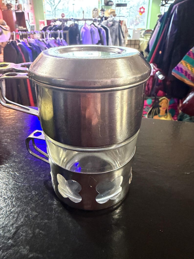 Puede incluir: Una cafetera vintage de acero inoxidable con una jarra de cristal. La cafetera tiene tapa, asa y una base decorativa con dise&ntilde;os recortados. La cafetera est&aacute; sobre una superficie oscura.