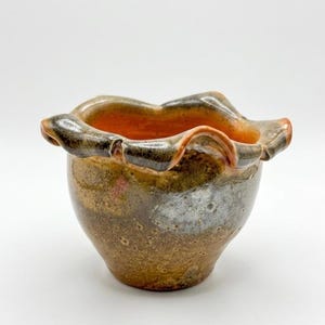 May include: A handcrafted ceramic bowl with a ruffled rim. The bowl features a blend of earthy tones, including brown, orange, and gray, with a glossy finish. The unique shape and color palette make it a decorative piece.
