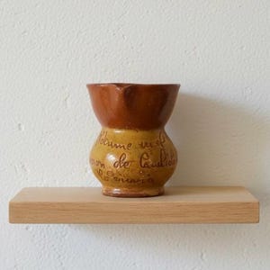 May include: A small, handcrafted ceramic vase with a two-tone design. The top is a rich brown, transitioning to a mustard yellow base. The vase features handwritten text and sits on a light-colored wooden shelf.