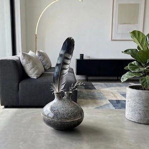 May include: A round, two-necked ceramic vase with a speckled black and white glaze. A large, dark, striped feather is placed in one of the openings, while a small, dried, light-colored twig is positioned in the other opening of the vase.