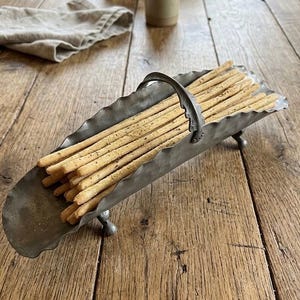 May include: A metal serving tray filled with breadsticks, set on a wooden table. The tray has a handle and decorative feet. The breadsticks are long, thin, and lightly seasoned. The table has a rustic, aged appearance.