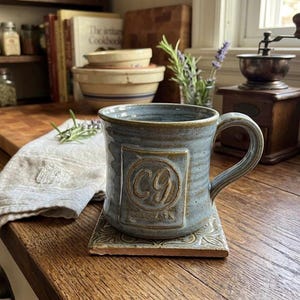 May include: A handmade blue ceramic mug with a handle, resting on a decorative tile coaster. The mug features the initials "CG" and the word "MACGAIX" in a rectangular design. The mug is on a wooden surface.