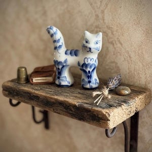 May include: A white ceramic cat figurine with blue floral patterns and stripes on its tail, standing on a rustic wooden shelf with a metal thimble, a small leather-bound book, a sprig of dried lavender, and a smooth pebble.