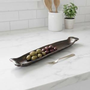 May include: A long, narrow, dark brown metallic serving tray with curved ends, containing a mix of green and dark purple olives. A small, slender silver-toned metal fork rests on the surface next to the tray, positioned to the right side.