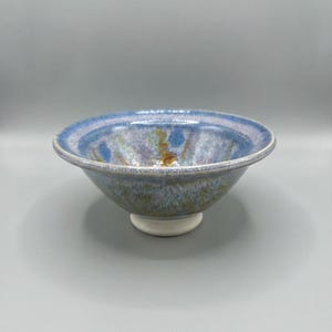 May include: A ceramic bowl featuring a wide, flared rim and a tapered base. The interior and exterior surfaces display a mottled glaze with blended shades of blue, lavender, and hints of brown, while the foot of the bowl is a solid white.