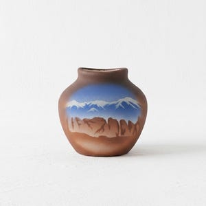 May include: A small, brown ceramic vase with a landscape design. The vase features a blue sky with white mountain peaks and a brown, rocky foreground. The vase has a rounded body and a narrow opening.