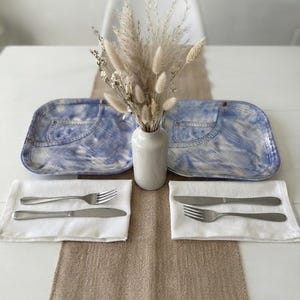 May include: Two square ceramic plates with a blue and white denim-inspired pattern, each featuring a pocket design. A white ceramic vase holds dried beige grasses between the plates. Two white cloth napkins with silver forks and knives.