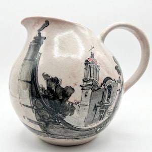 May include: A ceramic pitcher with a round body, a spout, and a handle. The pitcher is painted with a light pink background and a black and gray illustration of a building and trees. The pitcher has a crackled surface.