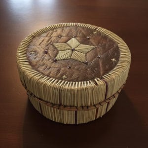 May include: A round, handcrafted box with a star design. The lid is brown leather with an embroidered star and small purple accents. The sides are made of woven, light-colored reeds, and the box is secured with a thin, brown band.